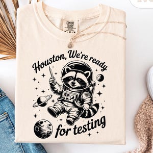 May include: Cream-colored t-shirt featuring a raccoon astronaut illustration with the text "Houston, We're ready for testing." The raccoon is wearing a spacesuit and holding a pen. The design includes planets and stars.