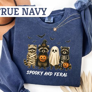 May include: Navy blue sweatshirt with a Halloween-themed graphic. The design features raccoons dressed as a mummy, witch, ghost, and skeleton, holding jack-o'-lanterns. The text "Spooky and Feral" is printed below the characters. The sweatshirt is labeled "True Navy".