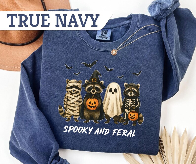 May include: Navy blue sweatshirt with a Halloween-themed graphic. The design features raccoons dressed as a mummy, witch, ghost, and skeleton, holding jack-o'-lanterns. The text "Spooky and Feral" is printed below the characters. The sweatshirt is labeled "True Navy".