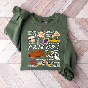 May include: An olive green sweatshirt featuring a graphic design with iconic imagery and quotes from the TV show "Friends." The design includes a couch, Central Perk logo, and phrases like "How you doin'?" and "Pivot!"