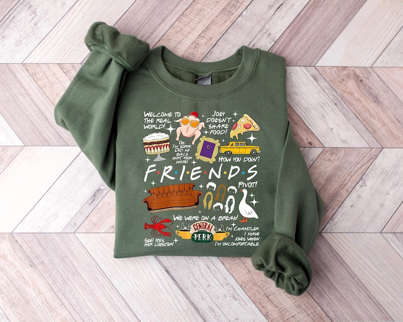 May include: An olive green sweatshirt featuring a graphic design with iconic imagery and quotes from the TV show "Friends." The design includes a couch, Central Perk logo, and phrases like "How you doin'?" and "Pivot!"