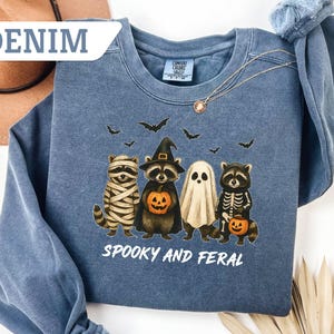 May include: Denim blue sweatshirt with a Halloween-themed graphic. The design features four cartoon raccoons dressed as a mummy, witch, ghost, and skeleton, holding jack-o'-lanterns. The text "Spooky and Feral" is printed below the characters.