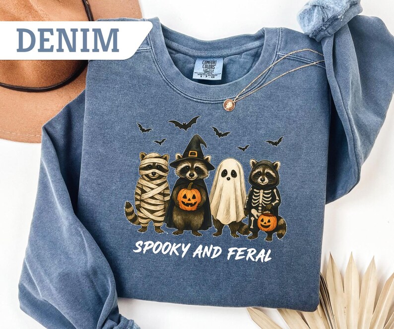 May include: Denim blue sweatshirt with a Halloween-themed graphic. The design features four cartoon raccoons dressed as a mummy, witch, ghost, and skeleton, holding jack-o'-lanterns. The text "Spooky and Feral" is printed below the characters.