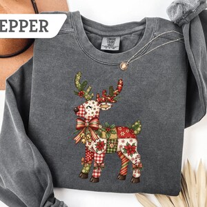 May include: A gray sweatshirt with a colorful patchwork reindeer design. The reindeer features a mix of patterns, including plaid, floral, and polka dots, with a large bow and festive accents. The word "PEPPER" is visible in the upper left corner.