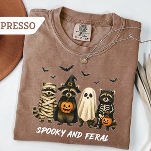 May include: Espresso-colored t-shirt with a Halloween-themed graphic. The design features four raccoons dressed as a mummy, a witch, a ghost, and a skeleton, holding jack-o'-lanterns. The text "Spooky and Feral" is printed below the raccoons.