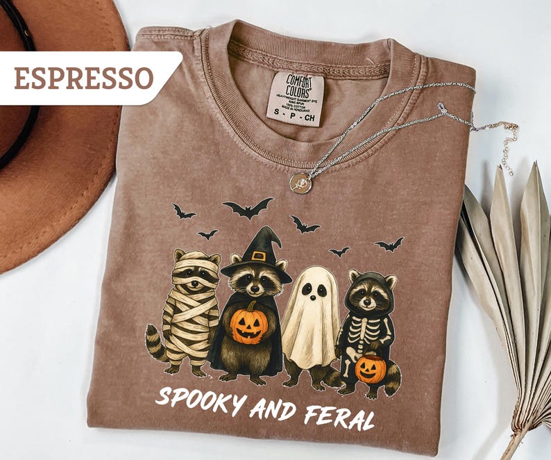 May include: Espresso-colored t-shirt with a Halloween-themed graphic. The design features four raccoons dressed as a mummy, a witch, a ghost, and a skeleton, holding jack-o'-lanterns. The text "Spooky and Feral" is printed below the raccoons.