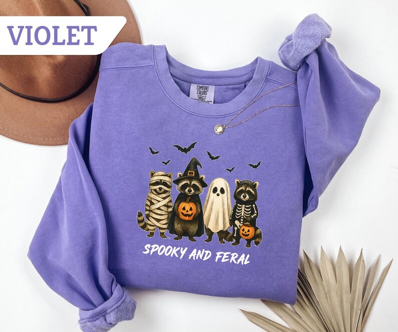 May include: Violet-colored sweatshirt featuring a Halloween-themed graphic. The design includes four raccoons dressed as a mummy, a witch, a ghost, and a skeleton, with bats flying above. The text "Spooky and Feral" is printed below the characters.