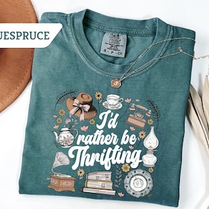 May include: Teal Comfort Colors t-shirt with the phrase "I'd rather be thrifting" and vintage-style illustrations of a hat, tea cup, telephone, and other antique items. A gold necklace is draped across the shirt.