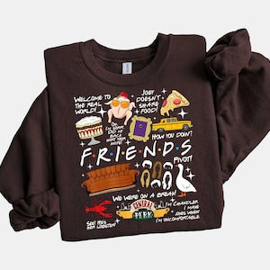 May include: A dark brown sweatshirt featuring various illustrations and text from the TV show "Friends." The design includes a turkey with a hat, a yellow taxi, a couch, and the Central Perk logo. Text includes "Welcome to the real world!" and "How you doin?"