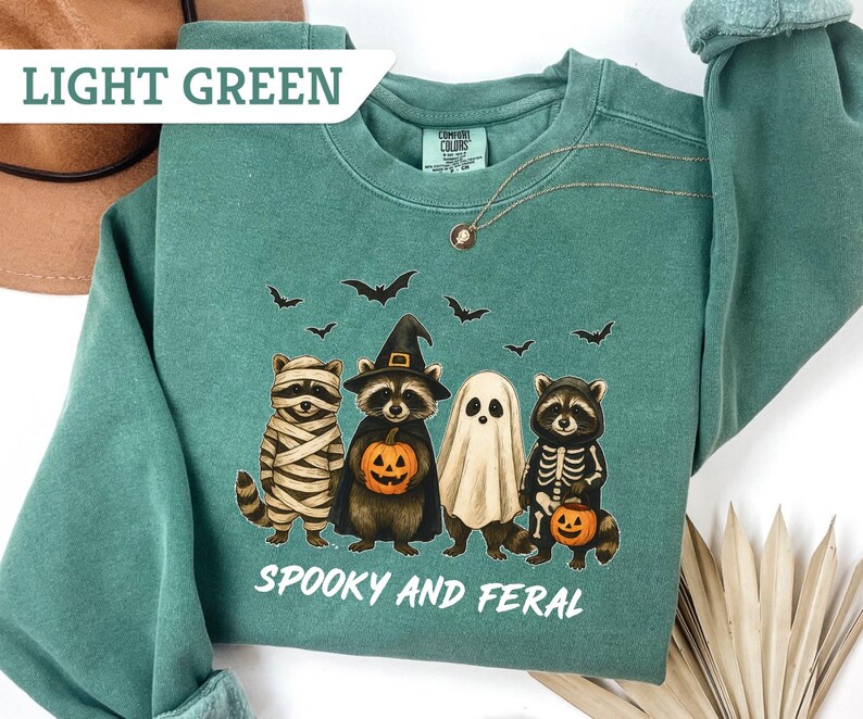 May include: Light green sweatshirt with a Halloween-themed graphic. The design features four raccoons dressed as a mummy, a witch, a ghost, and a skeleton, holding pumpkins. The text "Spooky and Feral" is printed below the raccoons.