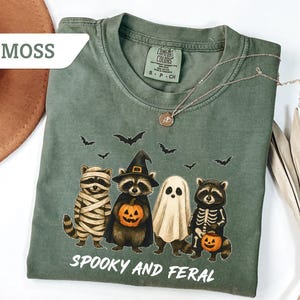May include: Moss green t-shirt featuring a Halloween-themed graphic. The design includes four raccoons dressed as a mummy, a witch, a ghost, and a skeleton, each holding a pumpkin. The text "Spooky and Feral" is printed below the illustration.