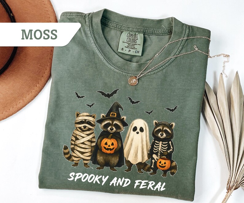 May include: Moss green t-shirt featuring a Halloween-themed graphic. The design includes four raccoons dressed as a mummy, a witch, a ghost, and a skeleton, each holding a pumpkin. The text "Spooky and Feral" is printed below the illustration.