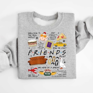 May include: A light gray sweatshirt featuring a colorful graphic design with text and illustrations from the TV show "Friends." The design includes the show's logo, a couch, and various quotes and images from the show.
