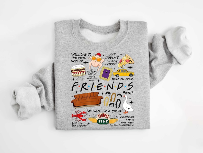 May include: A light gray sweatshirt featuring a colorful graphic design with text and illustrations from the TV show "Friends." The design includes the show's logo, a couch, and various quotes and images from the show.