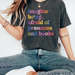 May include: Dark grey t-shirt with the text "imagine being afraid of pronouns and books" in colourful, retro-style lettering. The shirt is worn with blue jeans and white trainers. The overall theme is related to books and pronouns.