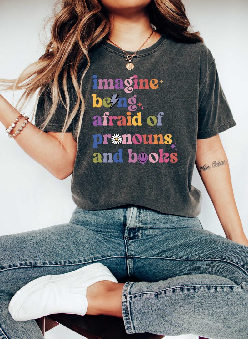 May include: Dark grey t-shirt with the text "imagine being afraid of pronouns and books" in colourful, retro-style lettering. The shirt is worn with blue jeans and white trainers. The overall theme is related to books and pronouns.