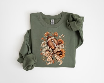 May include: Olive green sweatshirt featuring a watercolor design of a football surrounded by sunflowers, daisies, and a plaid bow. The long-sleeved top is a comfortable, casual style, perfect for fall and football season.