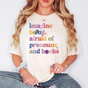 May include: A cream-coloured t-shirt featuring the phrase "imagine being afraid of pronouns and books" in a retro, multi-coloured font. The design includes a lightning bolt, a daisy, and a smiley face. The shirt is worn with blue jeans.