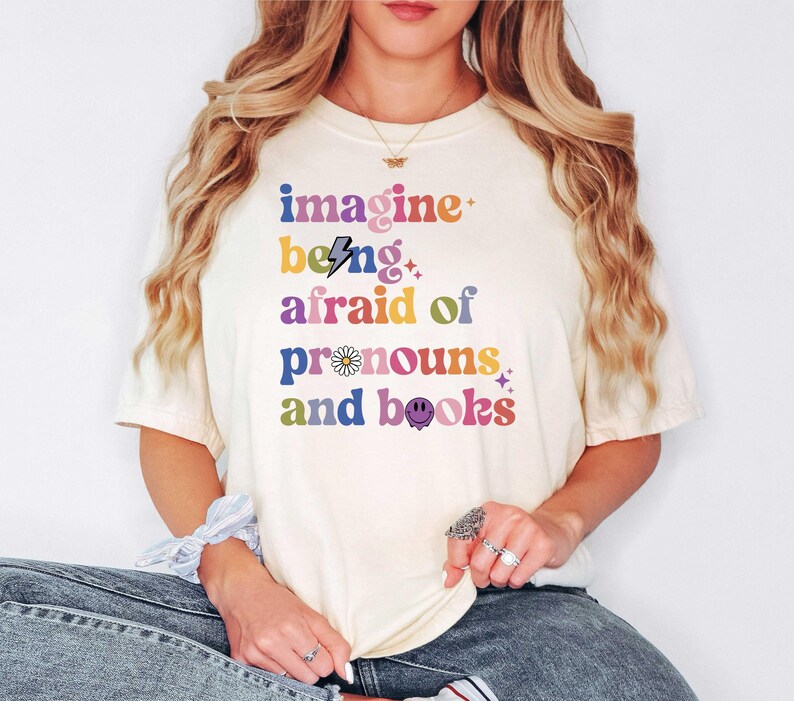 May include: A cream-coloured t-shirt featuring the phrase "imagine being afraid of pronouns and books" in a retro, multi-coloured font. The design includes a lightning bolt, a daisy, and a smiley face. The shirt is worn with blue jeans.