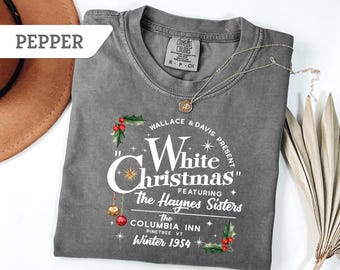 May include: A gray, crew neck t-shirt with the text "White Christmas" featuring The Haynes Sisters. The shirt has a vintage design with holly and ornaments. A silver necklace with a pendant is on the shirt.