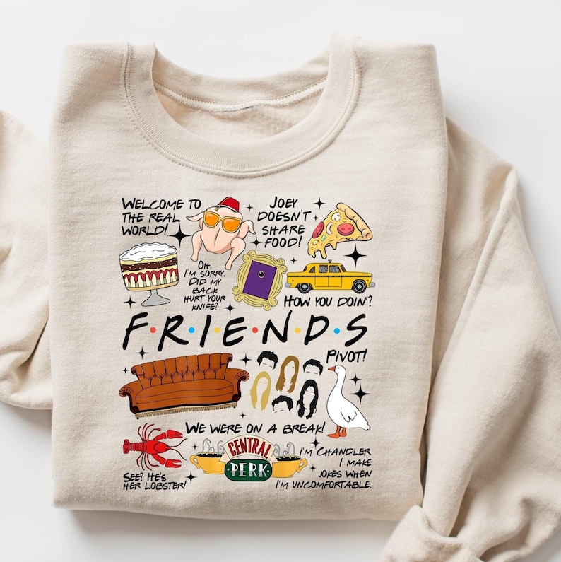 May include: Beige Friends-themed sweatshirt with various graphic elements. Features the show's logo and iconic imagery like a turkey with sunglasses, a yellow taxi, and the Central Perk logo. Text includes phrases from the show.