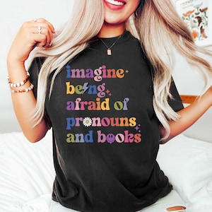 May include: Black t-shirt featuring the text "imagine being afraid of pronouns and books" in a vibrant, multi-coloured design. The graphic includes stars, a lightning bolt, and a daisy. The shirt is a relaxed fit.