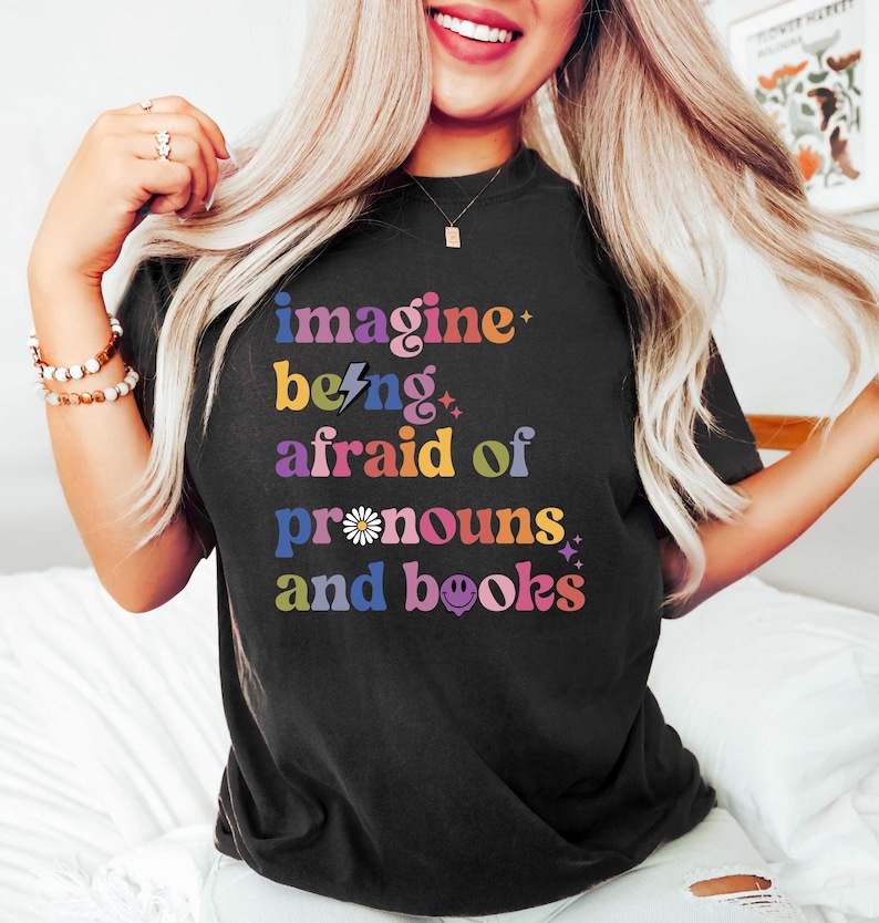May include: Black t-shirt featuring the text "imagine being afraid of pronouns and books" in a vibrant, multi-coloured design. The graphic includes stars, a lightning bolt, and a daisy. The shirt is a relaxed fit.