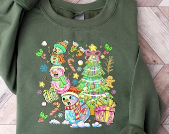 May include: A green sweatshirt featuring a colorful Christmas design. The graphic includes three snowmen wearing hats and scarves, a decorated Christmas tree, wrapped gifts, candy canes, and snowflakes. The design is festive and whimsical.