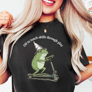 May include: Black t-shirt featuring a cartoon frog wearing a wizard hat and riding a scooter. The frog is green with a white hat. The text "Olf to teach skills through play" is arched above the frog.