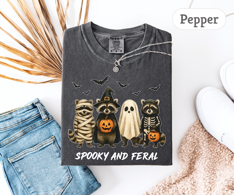 May include: Dark gray t-shirt with a Halloween-themed graphic. The design features four raccoons dressed as a mummy, a witch, a ghost, and a skeleton, holding pumpkins. The text "Spooky and Feral" is printed below the illustration.