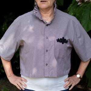 May include: A mauve and white striped, short-sleeved shirt with a ruffled collar and a black floral appliqué on the pocket. The shirt is cropped at the waist and has decorative stitching. The buttons are dark.