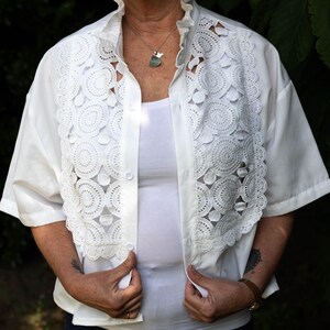 May include: White short-sleeved shirt with a decorative, openwork lace collar and front panel. The lace design features circular patterns. The shirt is buttoned and worn over a white vest top.