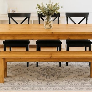 May include: A light brown wooden dining table with a matching bench and four black chairs. A clear glass vase with greenery sits on the table. The table is 72 inches long.