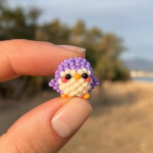 Puede incluir: Una pequeña figura de pingüino tridimensional hecha de diminutas cuentas de color morado, crema, negro, naranja y rosa. El pingüino tiene un cuerpo redondeado morado, un pecho color crema, ojos negros, un pequeño pico naranja y pies naranjas.