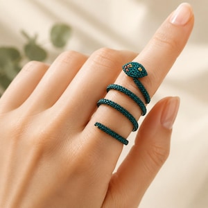 May include: A coiled snake ring crafted from dark teal seed beads. The design wraps around the finger in a spiral pattern, featuring a distinct head with small, multi-colored bead accents for eyes. The texture is uniform and granular throughout.
