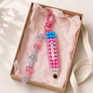 May include: A beaded keychain shaped like a pencil with a pink lobster clasp. The pencil body features pink beads with white letter beads spelling WASHU, blue top beads, and white bottom beads with a black tip. A dangling strand has pink and blue star beads.