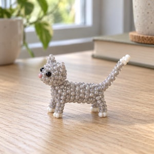 May include: A small cat figurine crafted from numerous tiny, shimmering silver-grey beads. The cat features black bead eyes, a small pink bead nose, and white beads on the tips of its four paws and its upright, pointed tail.