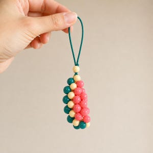 May include: A decorative charm featuring a cluster of small, round beads in shades of dark teal, cream, and pink. The beads are arranged in a curved, semi-circular shape and are suspended from a thin, dark teal cord loop for easy attachment.