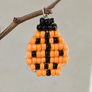 Könnte beinhalten: Ein Perlenanhänger in Form eines Marienkäfers, gefertigt aus kleinen orangefarbenen und schwarzen Perlen. Das Design zeigt einen zentralen schwarzen vertikalen Streifen mit symmetrischen schwarzen Punkten auf dem orangefarbenen Körper und einem schwarzen Kopf.