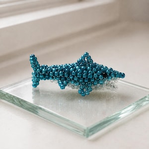 May include: A small shark figurine crafted from metallic blue beads with a white beaded underbelly. The shark features a prominent dorsal fin, side fins, and a tail fin, with a single dark bead serving as the eye on the side of the head.