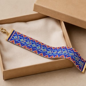 Op de afbeelding: Een brede kralenarmband met een geometrisch patroon in koningsblauw, feloranje en lichtturquoise. De armband heeft goudkleurige metalen eindstukken en een goudkleurige karabijnsluiting met een korte verlengketting.