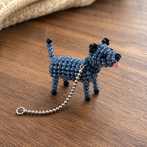 May include: A small dog figurine crafted from blue beads with black bead accents on the ears, tail, and paws. The dog features a pink bead tongue and a dark blue collar. A silver-toned metal ball chain leash with a circular ring is attached to the collar.