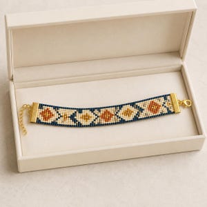 May include: A beaded bracelet featuring a geometric pattern in navy blue, cream, and burnt orange. The bracelet has gold-toned metal end caps and a lobster claw clasp, presented inside a cream-colored rectangular jewelry box with a hinged lid.