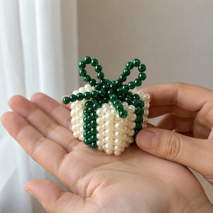 May include: A small, cube-shaped decorative object crafted from faux pearls. The main body consists of cream-colored pearls, while dark green pearls form a cross pattern and a bow on top, resembling a wrapped gift box with a decorative ribbon.