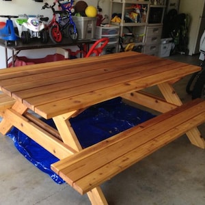 Könnte beinhalten: Ein Picknicktisch aus Holz mit angebrachten Bänken. Der Tisch ist aus hellbraunem Holz gefertigt und hat ein klassisches Design mit einer Lattenplatte und abgewinkelten Beinen. Der Tisch ist etwa 183 cm lang.