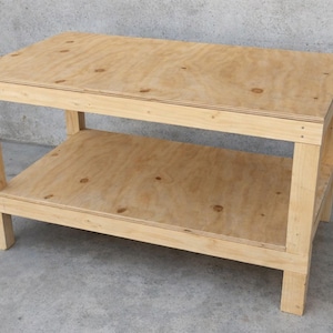 Puede incluir: Un banco de trabajo de madera sin terminar con una balda inferior. La mesa rectangular tiene cuatro patas y está construida con madera contrachapada. El banco de trabajo es adecuado para varios proyectos. Dimensiones: aprox. 120 cm de largo, 60 cm de ancho y 80 cm de alto.
