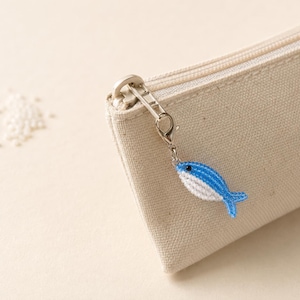 Op de afbeelding: Een beige canvas etui met een zilverkleurige metalen rits. Aan de ritssluiting hangt een kleine, handgemaakte bedel in de vorm van een vis, gemaakt van piepkleine blauwe en witte kraaltjes. Er liggen wat losse blauwe en witte kraaltjes bij.