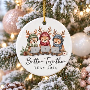 May include: A round white ceramic ornament featuring three illustrated reindeer wearing winter hats, scarves, and headphones while using laptops. The text reads "Better Together" and "TEAM 2026" with the names "EMMA", "OLIVIA", and "SOPHIA".