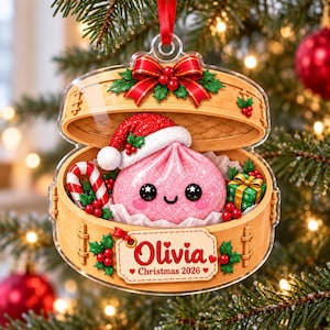 May include: A clear acrylic Christmas ornament shaped like a bamboo steamer basket containing a pink, glittery dumpling wearing a Santa hat. It features a candy cane, a green gift box, holly, and a tag reading Olivia Christmas 2026.