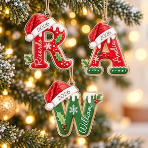 May include: Three hanging Christmas ornaments shaped like the letters R, A, and W. Each letter features a red Santa hat, white frosting, green holly leaves, red berries, and the year 2026. The letters are personalized with the names Ricardo, Amelia, and William.